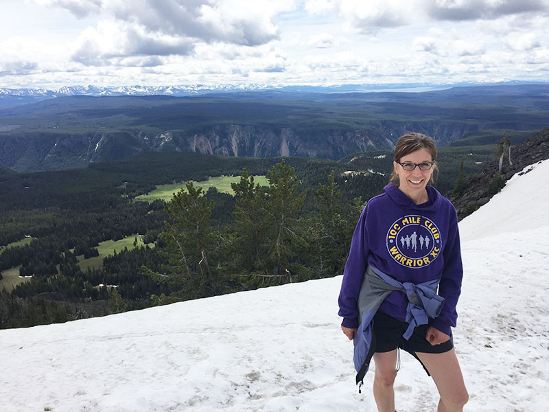 Dr. Sweet standing on a mountain in the snow wearing a purple sweatshirt with the words 100 mile club; trees and mountains in the background