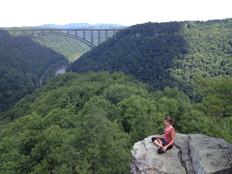 Woman sitting cross legged on stone ledge overlooking gorge with trees and bridge in background